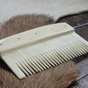Puede incluir: Un peine de hueso blanco con un mango decorativo. El mango tiene un patrón geométrico tallado y detalles dorados.