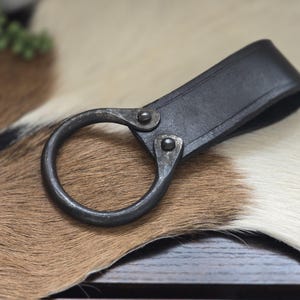 Puede incluir: Un llavero de cuero negro con un gran anillo metálico circular. La correa de cuero está remachada al anillo. El llavero se muestra sobre una superficie de piel de vaca marrón y blanca.