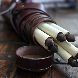 May include: A brown leather quiver containing rolled scrolls with dark brown tips. A matching leather container is in front. The items rest on a wooden surface, evoking a historical or fantasy setting.