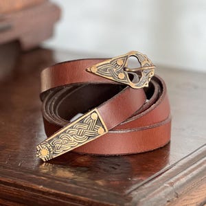 May include: A brown leather belt with a decorative gold-coloured buckle and belt end. The buckle and belt end feature an intricate Celtic knot design. The belt is coiled on a wooden surface, showcasing its craftsmanship and style.