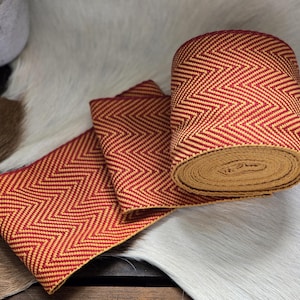 May include: A roll of red and yellow chevron patterned ribbon, with two flat pieces of the same ribbon. The ribbon is made of woven material and is displayed on a white and brown surface.