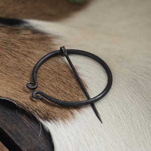 Puede incluir: Una broche de anillo de metal negro con un pasador. La broche est&aacute; sobre una superficie de piel blanca y marr&oacute;n.