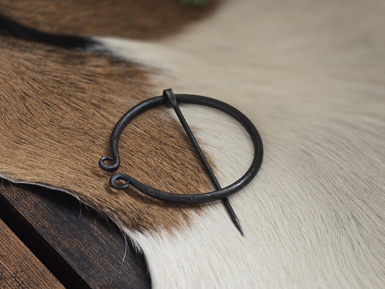 Puede incluir: Una broche de anillo de metal negro con un pasador. La broche est&aacute; sobre una superficie de piel blanca y marr&oacute;n.