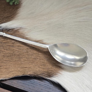 May include: A silver spoon with a long, slender handle and a rounded bowl. The handle has a decorative end. The spoon is lying on a brown and white patterned surface.