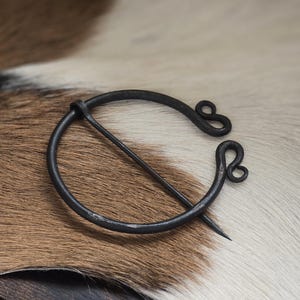 Puede incluir: Un broche de metal negro con un dise&ntilde;o circular y dos lazos decorativos. El broche est&aacute; sobre una superficie de piel marr&oacute;n y blanca.