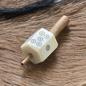 May include: A spinning top toy featuring a central off-white hexagonal die-shaped body with black circular markings on each face, mounted on a thin, cylindrical wooden spindle that extends through the center of the die to form the handle.