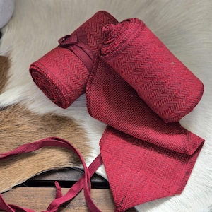 May include: Rolled red fabric with a herringbone pattern, tied with a matching ribbon. The fabric is arranged on a surface with a cowhide rug and a wooden plank visible in the background. The fabric is a deep shade of red.