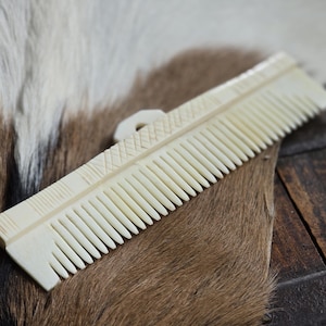 May include: A white bone comb with a decorative pattern on the handle. The comb has fine teeth and is lying on a brown and white fur surface.