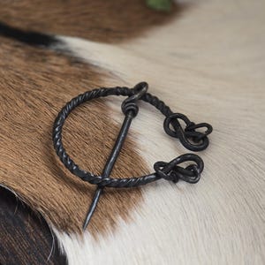 Puede incluir: Un broche de estilo vikingo de metal negro con un dise&ntilde;o de alambre retorcido y un pasador. El broche est&aacute; sobre una superficie de piel blanca y marr&oacute;n.
