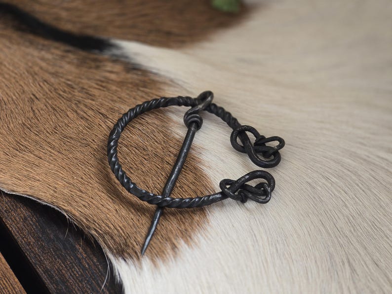 Puede incluir: Un broche de estilo vikingo de metal negro con un dise&ntilde;o de alambre retorcido y un pasador. El broche est&aacute; sobre una superficie de piel blanca y marr&oacute;n.