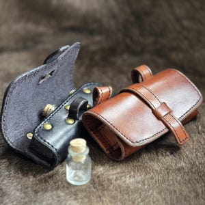 Może przedstawiać: Trzy skórzane sakiewki w kolorze czarnym i brązowym, z małą szklaną butelką z korkiem. Czarna sakiewka jest otwarta, odsłaniając korek. Brązowa sakiewka jest zamknięta paskiem. Sakiewki leżą na brązowej, teksturowanej powierzchni.