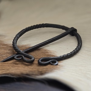 May include: A black metal Viking-style brooch with a twisted wire design and a long pin. The brooch is lying on a brown and white fur surface.