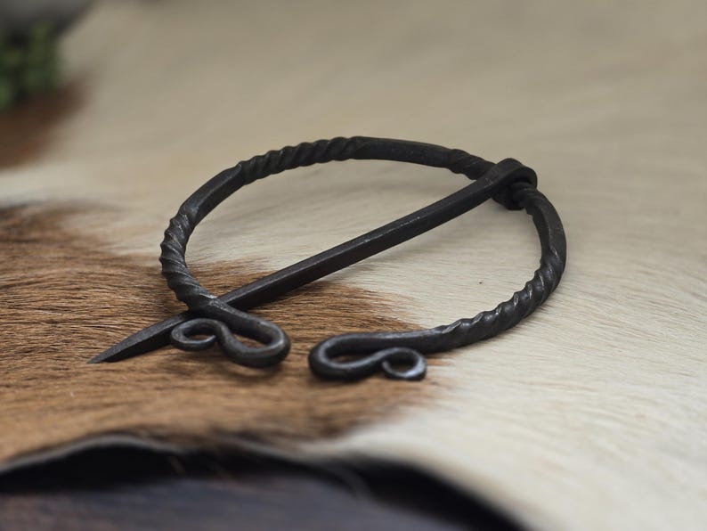 Puede incluir: Un broche de estilo vikingo de metal negro con un dise&ntilde;o de alambre retorcido y un pasador largo. El broche est&aacute; sobre una superficie de piel marr&oacute;n y blanca.