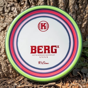 May include: A multi-colored disc golf disc with a white center and the text "BERG X" in red. The disc has a green outer rim, with pink, red, and blue rings. The disc is set against a natural wood background.