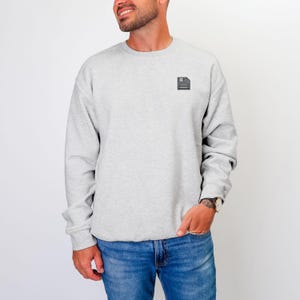May include: A light gray sweatshirt with a small black and gray floppy disk icon on the upper left chest. The sweatshirt has long sleeves and a crew neck. The person in the image is wearing blue jeans and a watch.