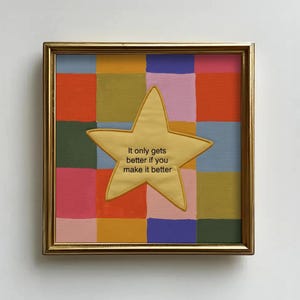 May include: A framed artwork with a gold border. The artwork features a colorful patchwork background of squares in various colors. A gold star with the text "It only gets better if you make it better" is centered on the artwork.