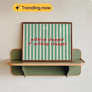 May include: A framed print with a brown border, featuring vertical green and white stripes and the text "nothing changes if nothing changes" in red. The print rests on a two-tiered, green and beige shelf, mounted on a white wall. A yellow banner with the text "Trending now" is in the top left corner.