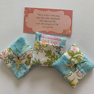 May include: Three small, quilted fabric pouches with a floral pattern and a gold cross charm. The pouches are in shades of blue, pink, and white. A card with text from John 6:47-48 is in the background.