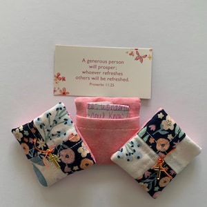 May include: An arrangement featuring two small, floral-patterned fabric pouches with gold cross charms, a central pink pouch, and a card with the text "A generous person will prosper; whoever refreshes others will be refreshed. Proverbs 11:25."