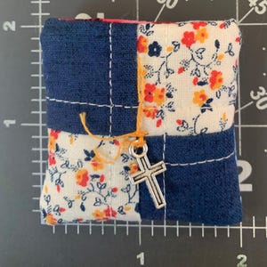 May include: A small, square fabric item with a patchwork design. It features sections of dark blue denim and floral fabric with red and yellow flowers on a white background. A silver cross charm hangs from an orange thread. The item is approximately 1.5 inches square.