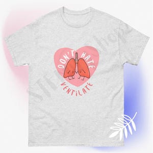 Op de afbeelding: Een grijs T-shirt met een roze hartontwerp. Het hart bevat een paar cartoonlongen met de tekst "Don't Hate Ventilate" rond het hart.