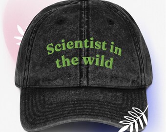 May include: A black, distressed baseball cap with the embroidered text "Scientist in the wild" in green. The cap has a curved brim and a classic six-panel design, suitable for outdoor activities or casual wear.