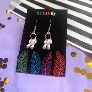 Peut inclure: Boucles d'oreilles en forme d'astronaute blanches avec des plaques faciales noires et un crochet argenté. Les boucles d'oreilles sont présentées sur une carte noire avec le texte "STEM-ily" imprimé dessus.