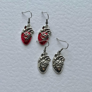May include: Two pairs of anatomical heart earrings with silver-tone metal hooks. One pair features a vibrant red enamel finish on the heart-shaped pendants, while the other pair consists of solid silver-tone metal pendants with detailed textures.
