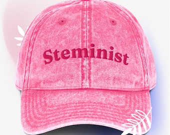 May include: A pink baseball cap with the word "Steminist" embroidered in a darker pink. The cap has a curved brim and a small, white leaf design on the side. The hat is made of a soft, washed fabric.
