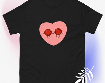 May include: Black t-shirt featuring a pink heart with two red, round characters holding hands. The characters have small legs and are smiling. The design is centered on the shirt.