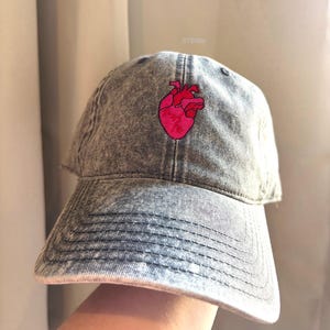May include: A grey denim baseball cap featuring a pink embroidered anatomical heart. The cap has a curved peak and an adjustable strap. The text "STEMily" is visible on the top right of the cap.