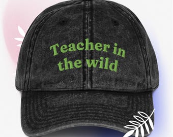May include: A black, distressed baseball cap with the text "Teacher in the wild" embroidered in green. The cap has a curved brim and a black button on top. A white leaf design is on the lower right side.