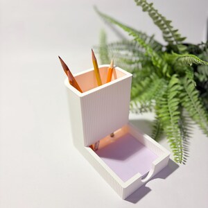 May include: A white desk organizer with a ribbed texture, holding several pencils. The organizer has a pull-out drawer with a purple interior. A green plant is in the background.