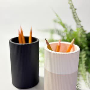 May include: Two cylindrical pencil holders, one black and one cream and white, each filled with sharpened pencils. The holders have a ribbed texture and are set against a white background with greenery in the distance.