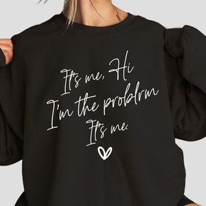 May include: Black sweatshirt with white text that reads "It's me. Hi I'm the problem It's me." and a small heart.