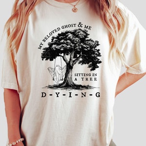 May include: A white t-shirt with a black and white illustration of a ghost sitting on a swing in a tree. The text "MY BELOVED GHOST & ME" is above the tree and the text "SITTING IN A TREE" is below the tree. The text "D-Y-I-N-G" is below the tree.