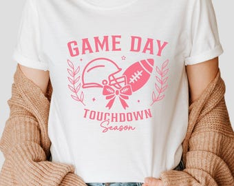 May include: White t-shirt with a pink graphic design that says "Game Day Touchdown Season". The graphic features a football helmet and football with a pink bow.