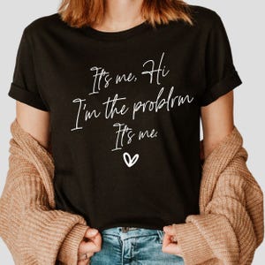 May include: Black t-shirt with white text that reads "It's me. Hi I'm the problem It's me." and a small white heart.