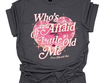 May include: A dark gray t-shirt with a pink glitter heart design. The text on the shirt reads "Who's Afraid of Little Old Me (You Should Be)".