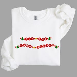 May include: White sweatshirt with the text "Tis the Damn Season" in red circles with green Christmas trees on either side of the text.