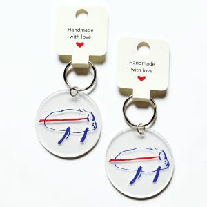 May include: Two circular clear acrylic keychains featuring a blue line drawing of a creature with a horizontal red line across its body. Each keychain is attached to a white card with the text Handmade with love and a small red heart icon.
