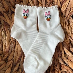 May include: A pair of white ribbed ankle socks featuring a ruffled top edge and an embroidered graphic of Daisy Duck on the front. The character design includes a pink bow, blue eyelids, and an orange beak, centred on the upper part of each sock.