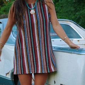 May include: A sleeveless crochet mini dress featuring vertical stripes in a vibrant multicolored pattern of red, blue, black, white, and orange. The dress has a high neckline and a straight hemline, crafted with a textured, open-knit stitch.