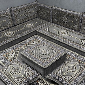 Puede incluir: Un conjunto de asientos de suelo de color gris y blanco con un gran sofá en forma de L, cuatro cojines cuadrados y un pequeño puf cuadrado. El conjunto está hecho de tela y tiene un diseño tradicional.