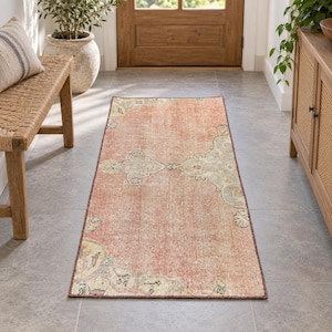May include: A rectangular area rug featuring a faded coral-orange center with intricate, muted sage green and cream-colored floral patterns along the borders and corners, laid flat on a gray tiled floor in a hallway setting.