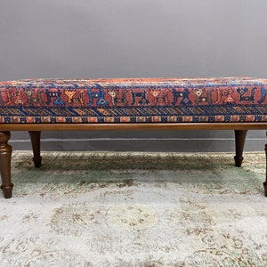 May include: A wooden bench with a patterned fabric seat cushion. The fabric has a red, blue and orange geometric design. The bench has four turned legs.