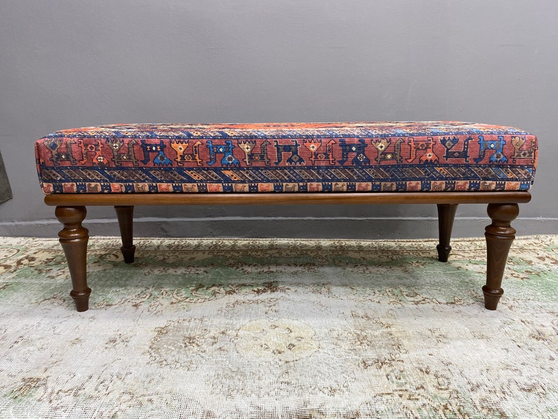 May include: A wooden bench with a patterned fabric seat cushion. The fabric has a red, blue and orange geometric design. The bench has four turned legs.