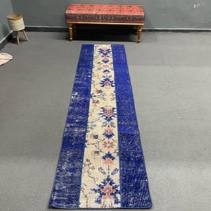 Pode incluir: Um tapete longo e retangular com um painel central creme com padrões florais em rosa, azul e verde. As laterais são azuis escuros, criando uma borda contrastante. O tapete está colocado sobre um chão cinzento.