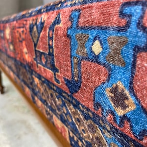 May include: A close-up of a wooden bench with a red, blue and brown patterned fabric seat. The fabric has a traditional design with geometric shapes and floral motifs.