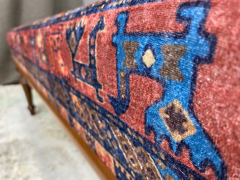May include: A close-up of a wooden bench with a red, blue and brown patterned fabric seat. The fabric has a traditional design with geometric shapes and floral motifs.